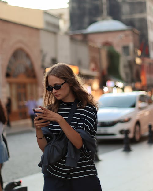 A Woman in a Striped Shirt Using a Smartphone on a Street