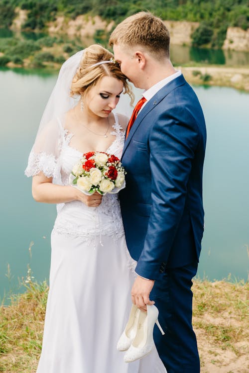 Novio Sosteniendo Un Par De Zapatos De Tacón De Cuero Blanco De Pie Delante De La Novia En Vestido De Novia Blanco