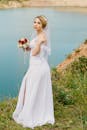 美丽的新娘身着白色连衣裙，手捧红白相间的花束，在碧绿的湖边摆姿势