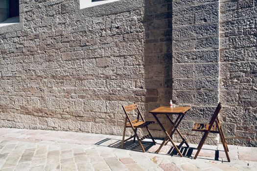 A sunny outdoor cafe scene with wooden chairs and table against a stone wall in Kotor, Montenegro.