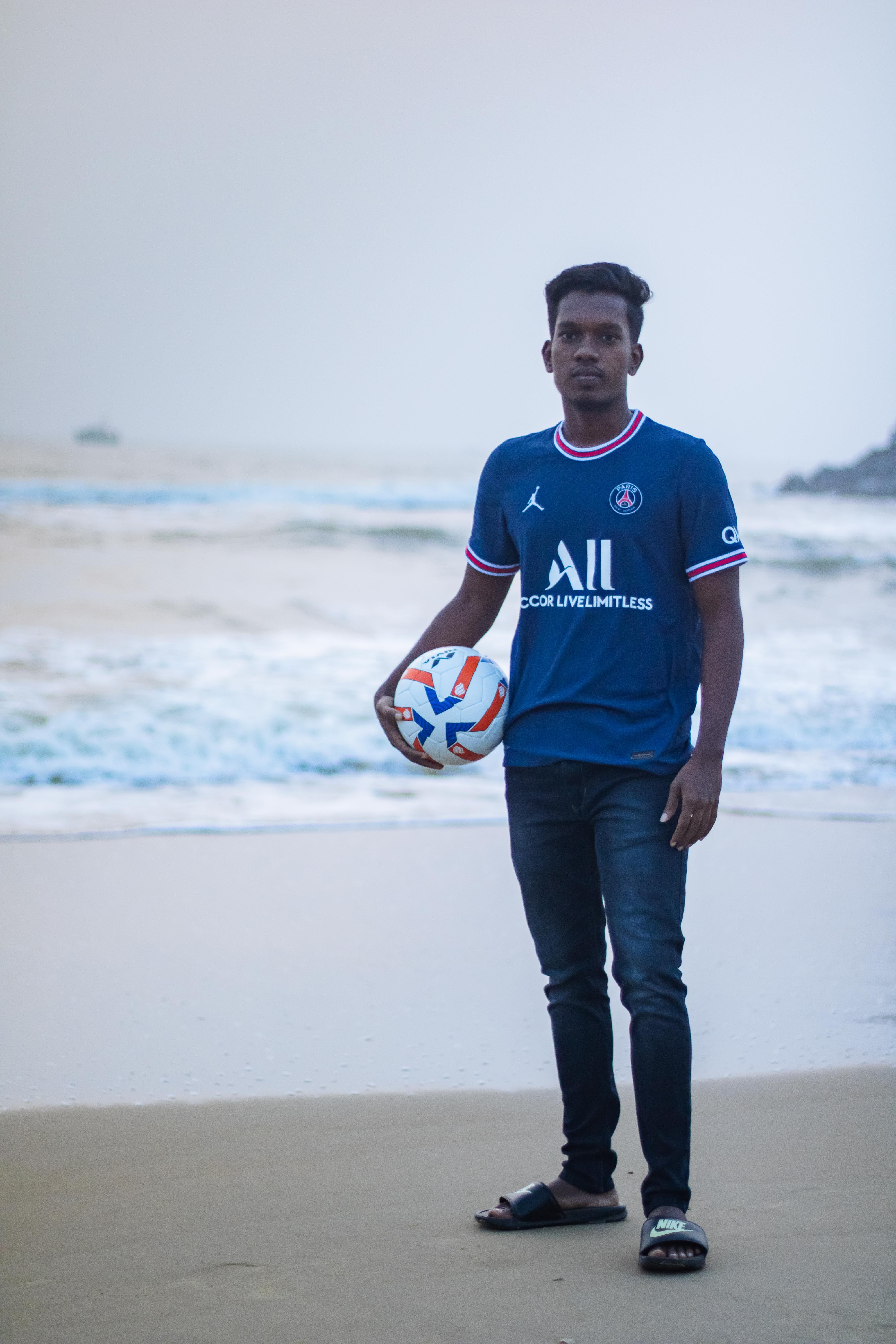 A young man stands on a beach holding a soccer ball with the sea in the background.
