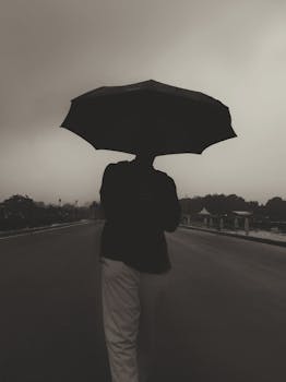 Black and white silhouette of a person holding an umbrella on a rainy street, creating a moody atmosphere.