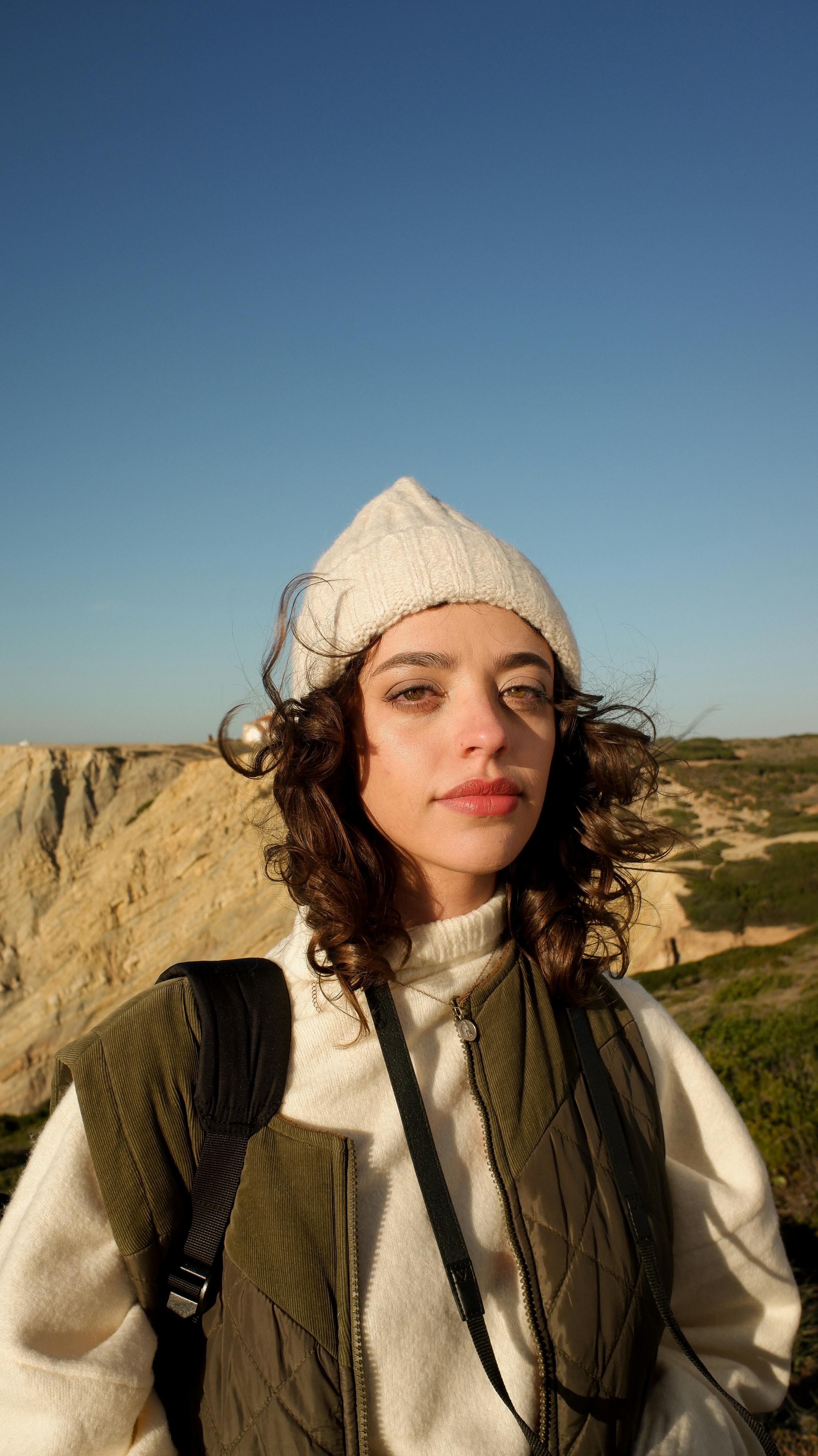 Young woman wearing a beanie, standing outdoors with a mountainous landscape, exuding a sense of adventure.