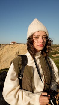 A young woman in a beanie and puffer vest holds a camera on a sunny cliffside.