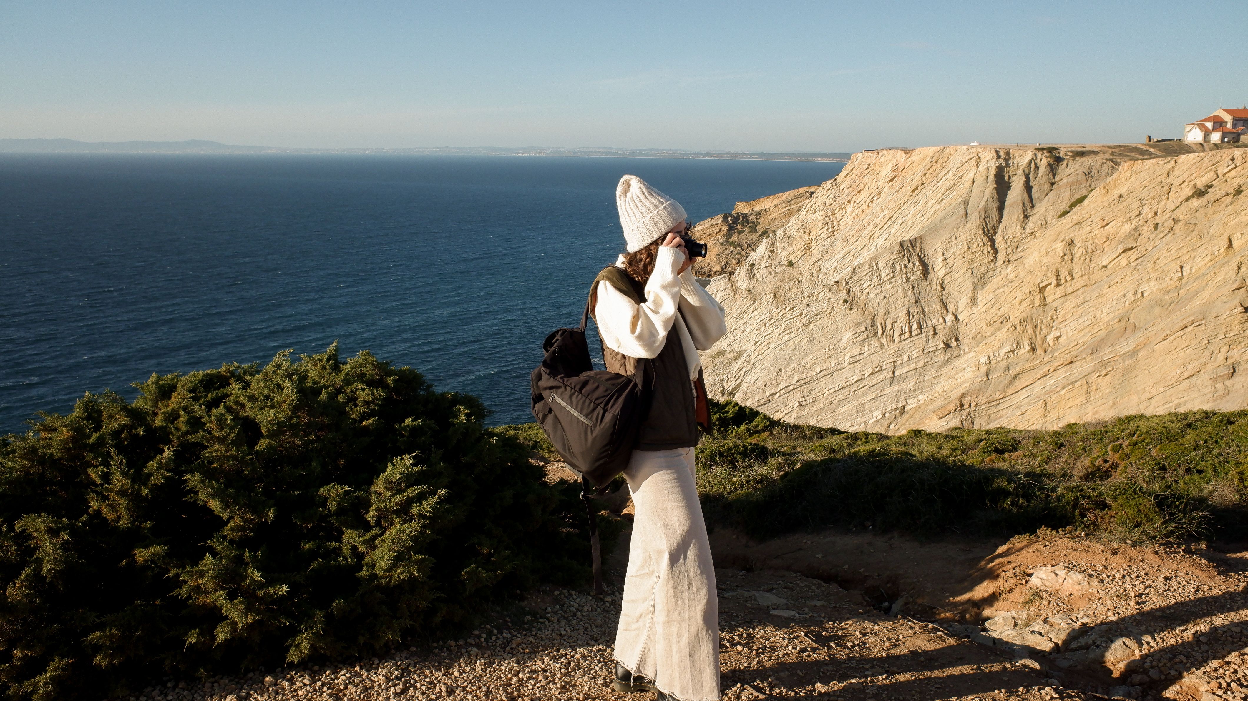 Woman in warm attire photographing a breathtaking ocean cliff during golden hour.