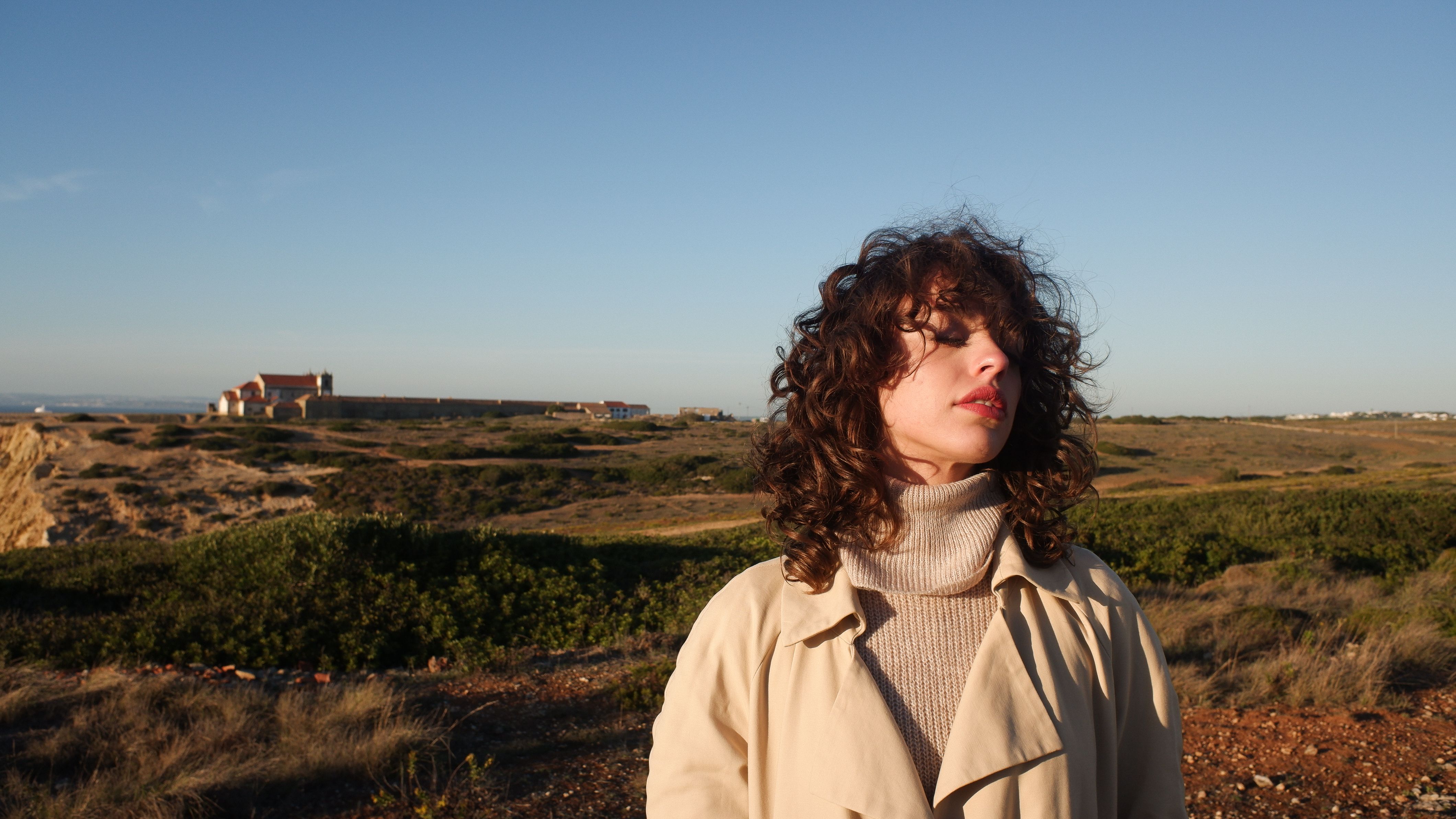Woman in a coat standing outdoors near a lighthouse enjoying a sunset in the countryside.
