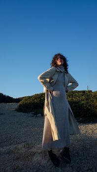 A fashionable woman poses in a trench coat against a blue sky with shrubs in the background.