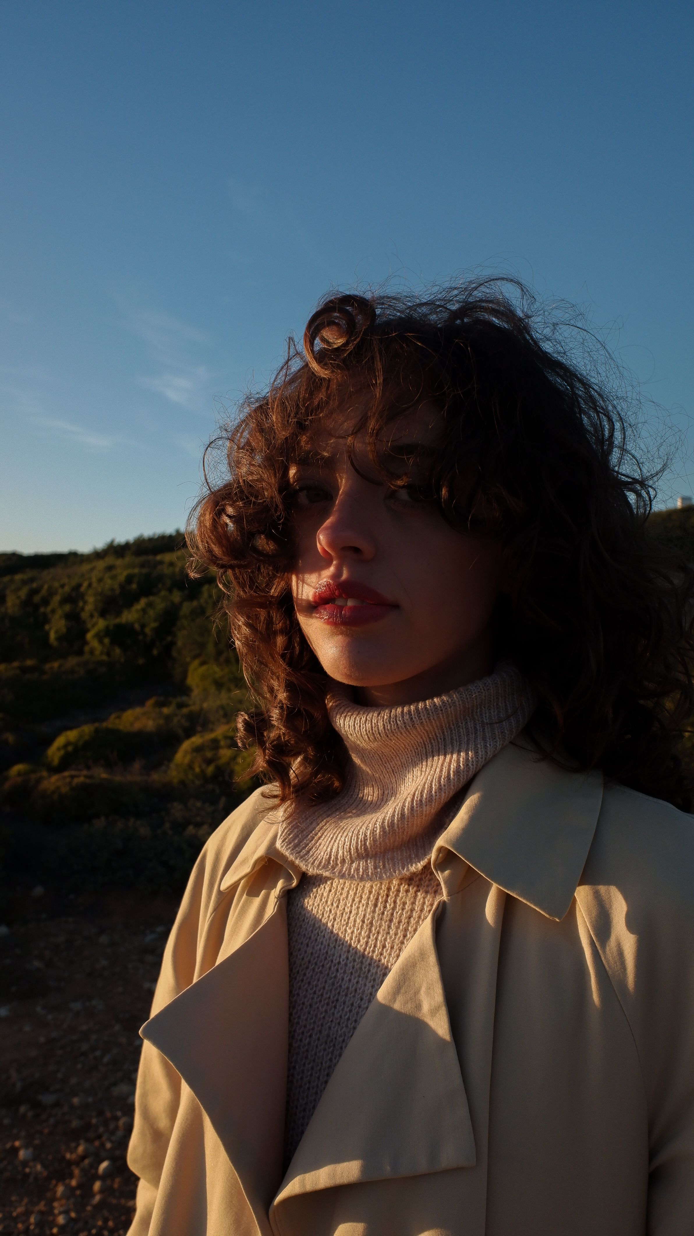 A woman with curly hair poses outdoors in a beige coat and turtleneck during sunset.