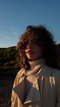A woman with curly hair poses outdoors in a beige coat and turtleneck during sunset.