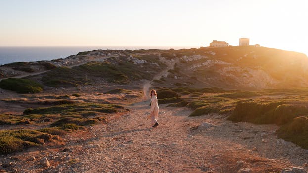 A woman strolls on a rocky coastal path during sunset, with a distant house on the horizon.