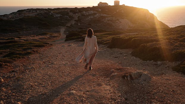 A woman walks along a coastal cliff path during golden hour, with the dramatic sunset in the background.