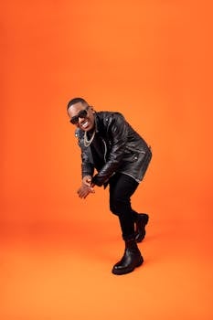 Fashion model wearing a leather jacket and boots posing dynamically against an orange studio background.