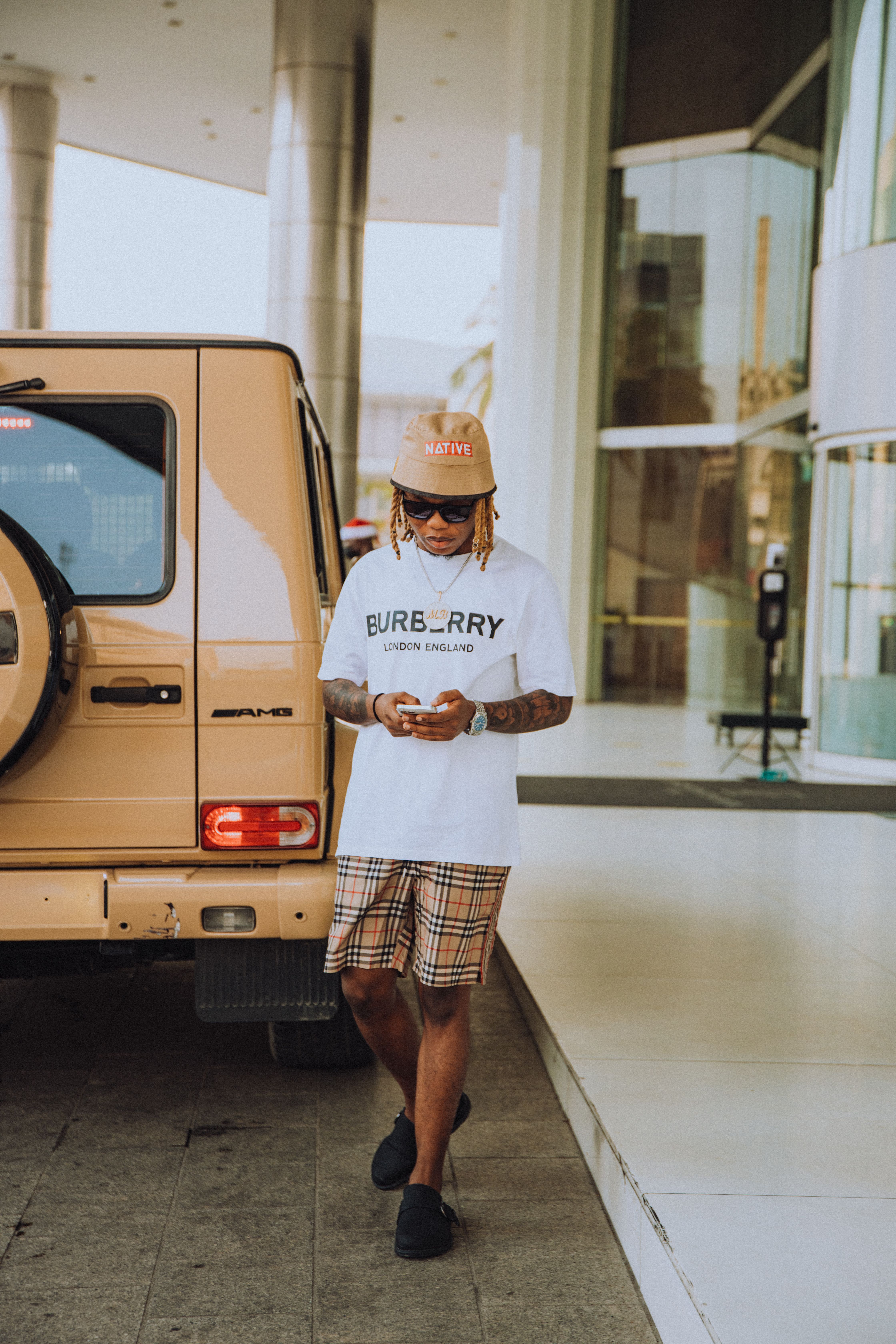 A fashionable man with afro braids and sunglasses checks his phone next to a luxury SUV.