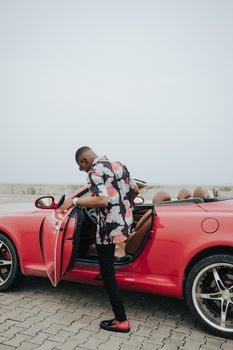 A man in a floral shirt opens the door of a red luxury convertible parked outdoors.