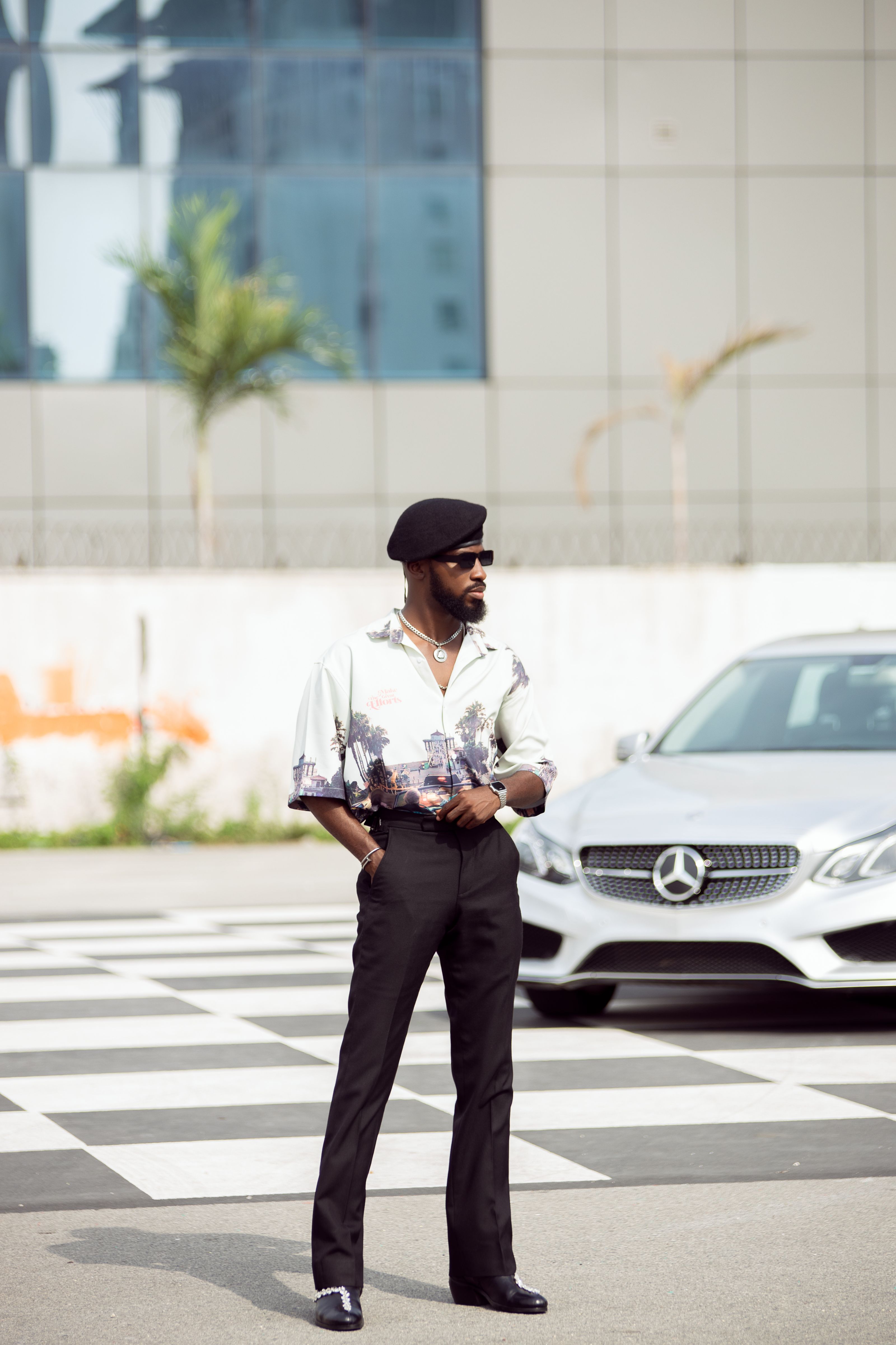 Homem elegante posando confiantemente em uma rua da cidade com um carro Mercedes-Benz esguio ao fundo.