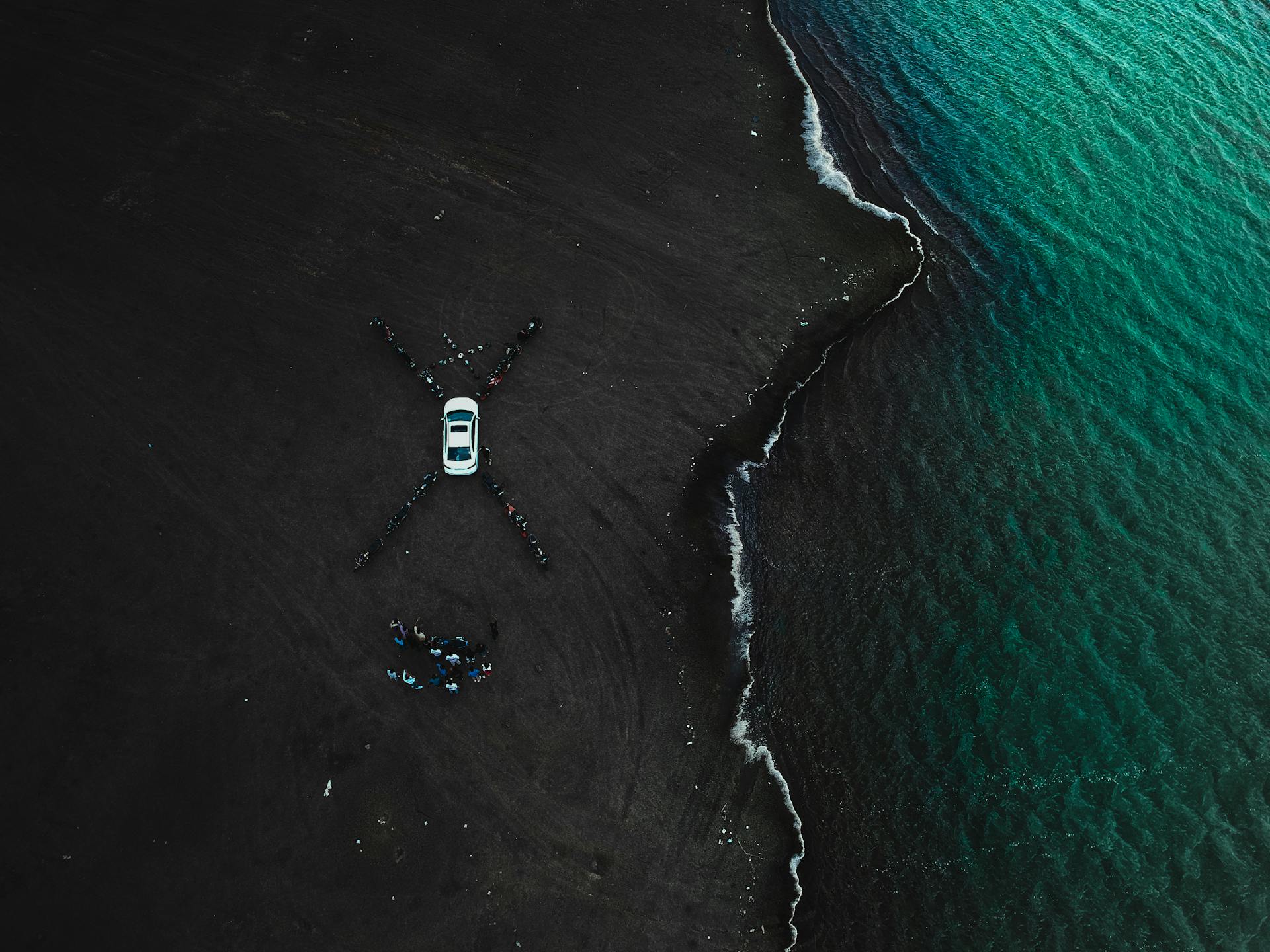 Drone shot capturing a car on a black sand beach by teal ocean waves.