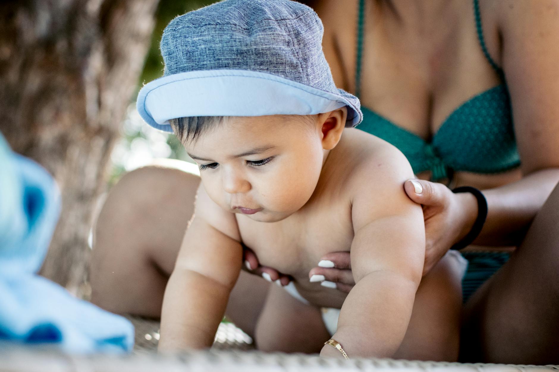 Adorable baby in a hat crawling under mother's supervision in a sunny outdoor setting.