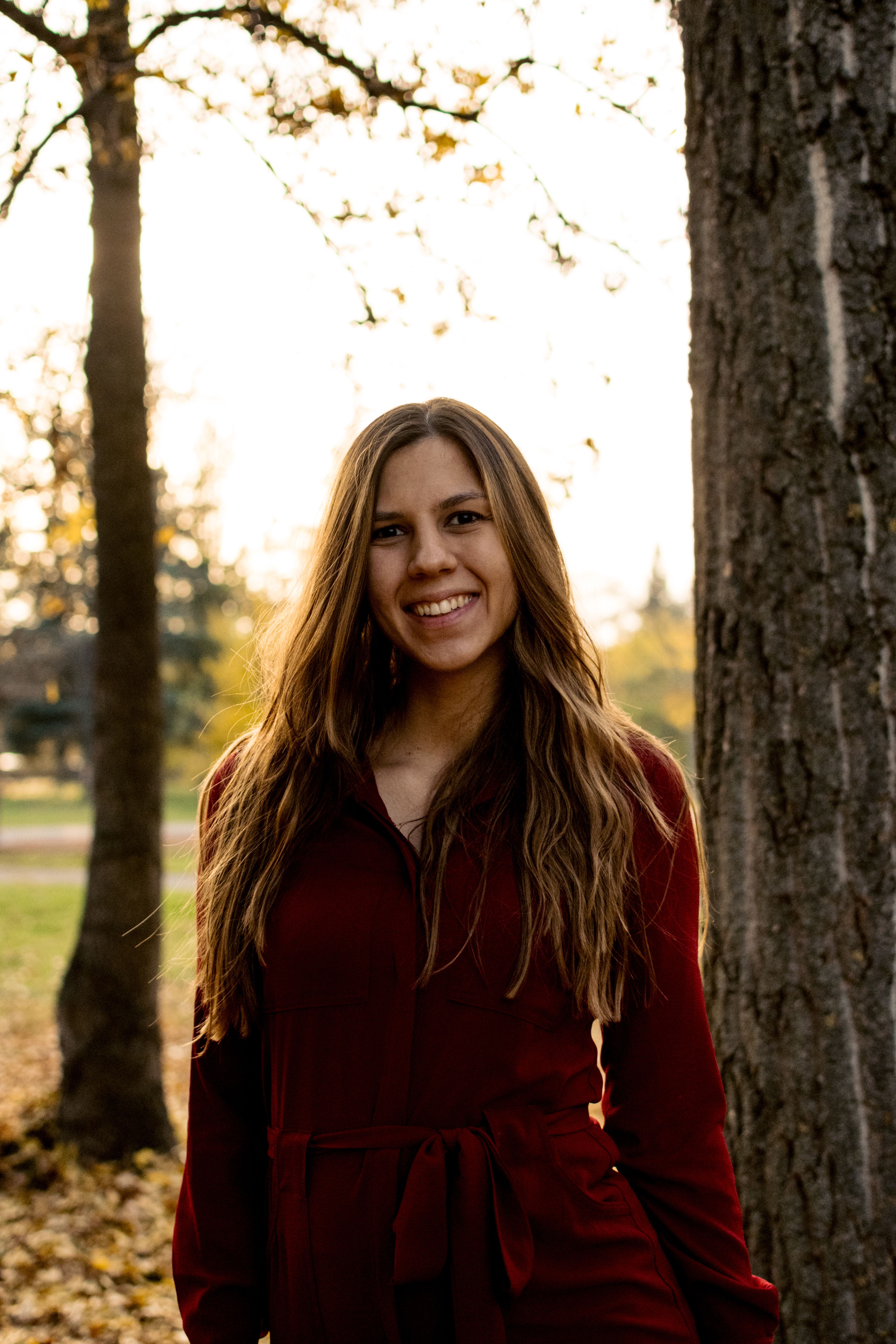A cheerful young woman in a red dress standing outdoors in autumn sunlight, surrounded by trees.