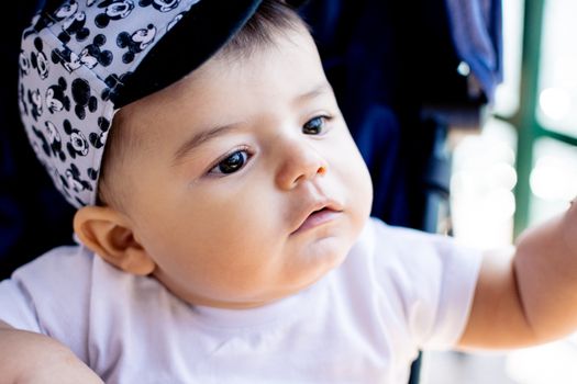 Close-up of a cute baby wearing a Mickey Mouse cap and white t-shirt, looking curious.