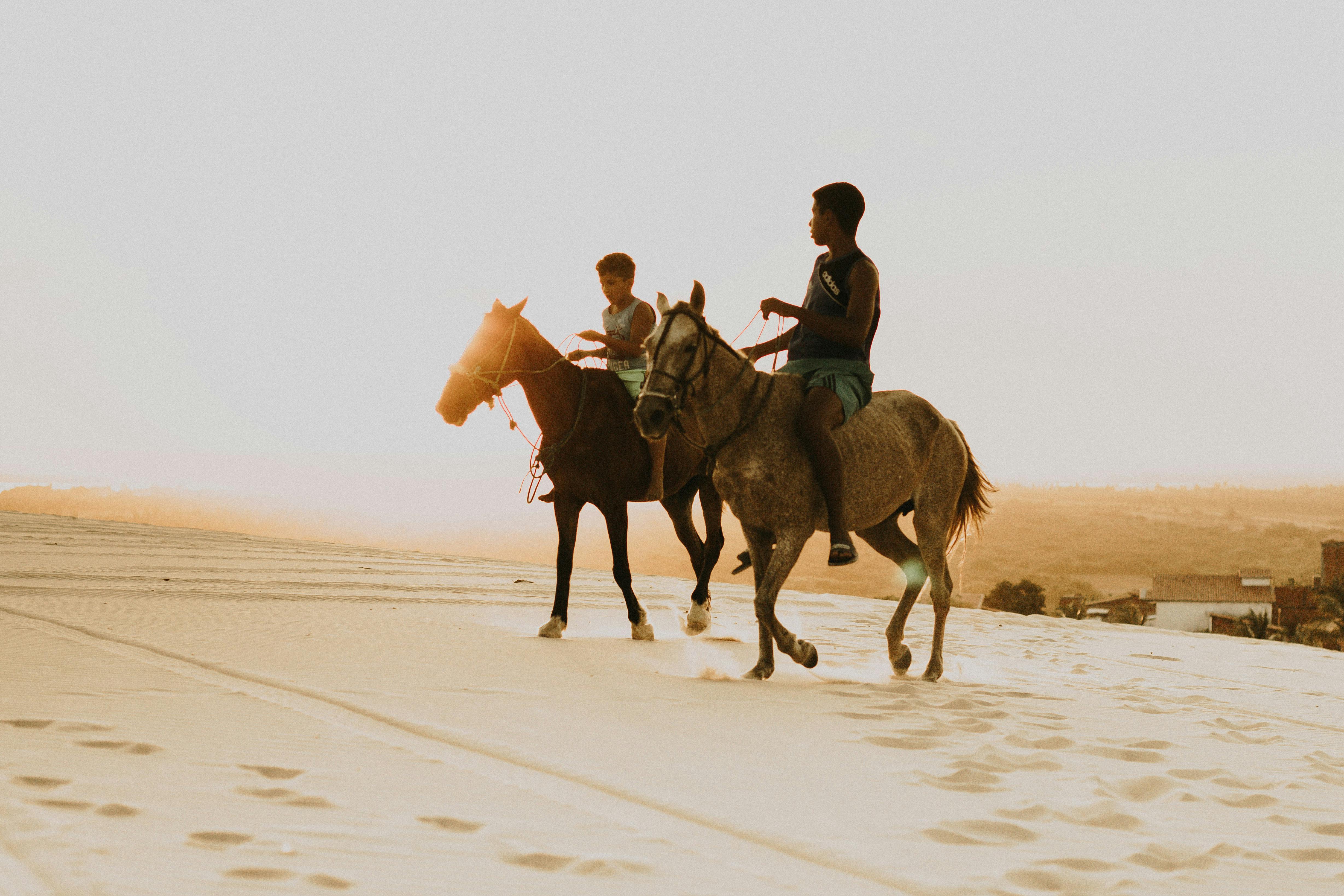 Two boys riding horses on the sandy dunes of Canoa Quebrada during sunset.