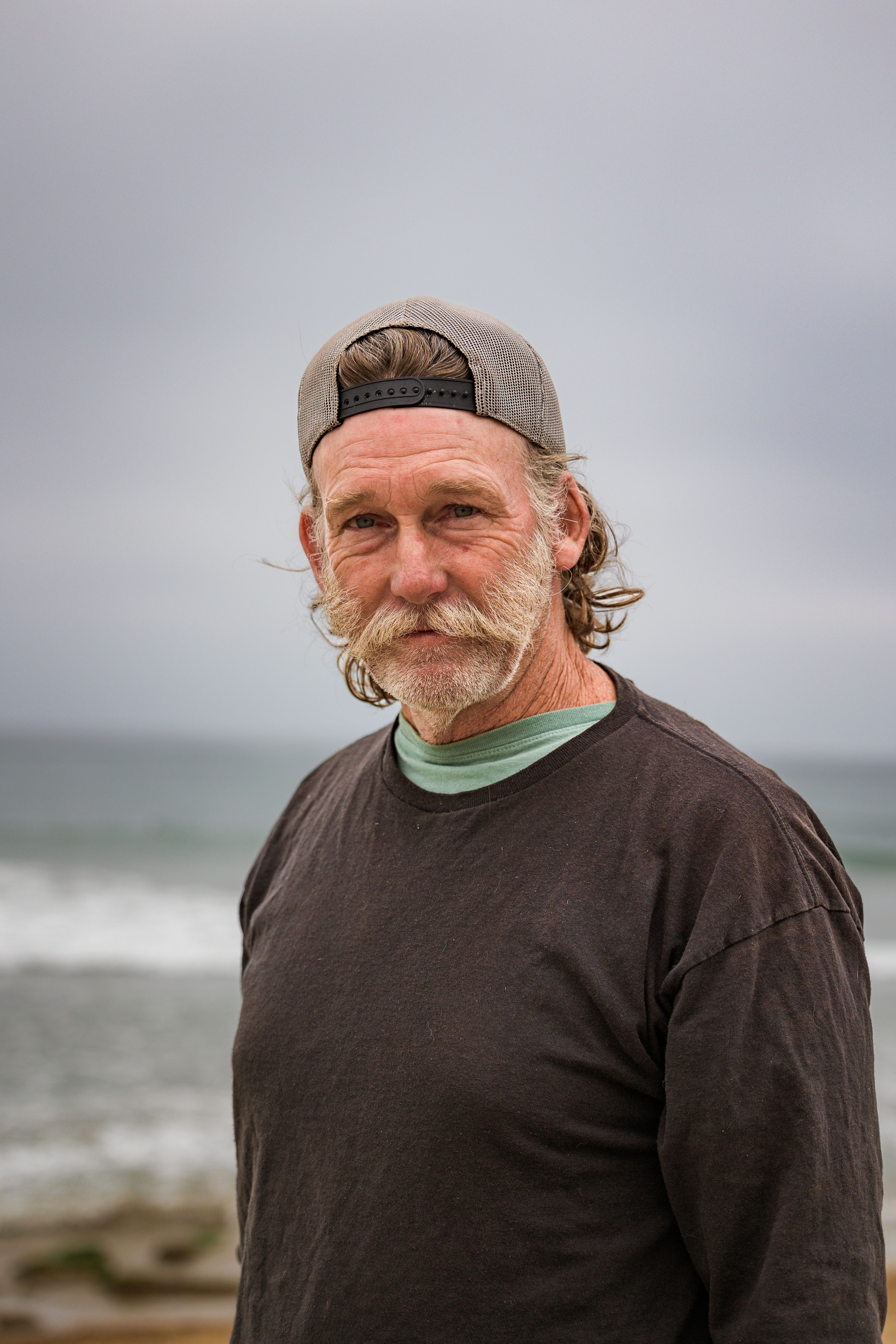 Elderly man with a beard and cap standing near the ocean shoreline.