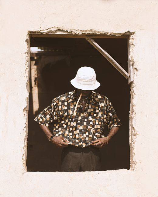 A stylish black man stands in a window frame wearing a patterned shirt and bucket hat.
