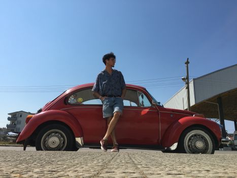 Man in denim shorts leaning on a vintage red car under blue sky.
