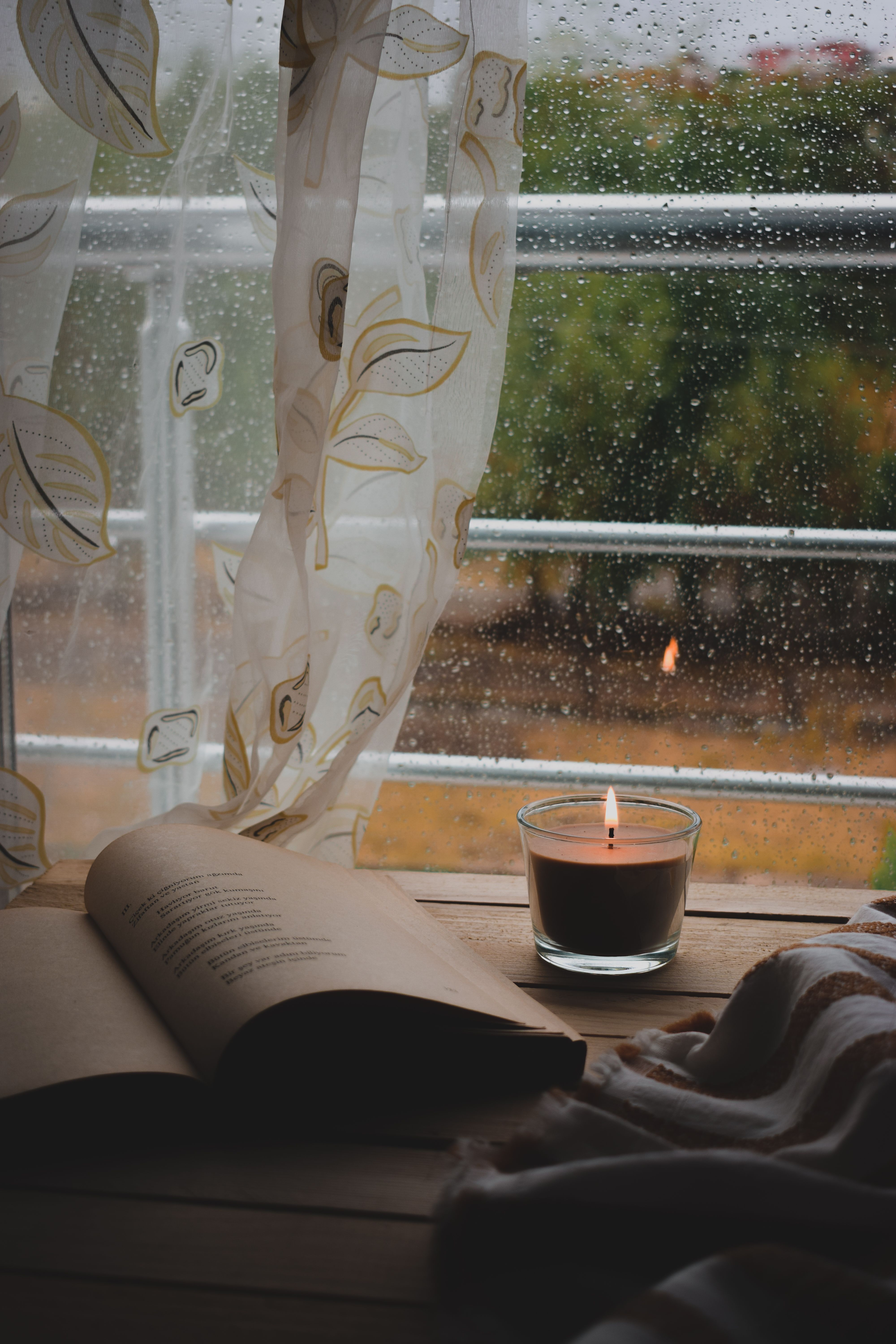 Open book and lit candle by a window with rain. Perfect for cozy reading ambience.