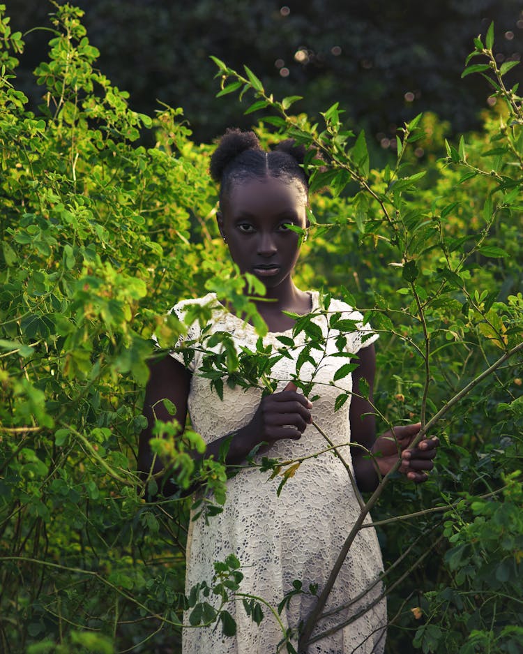 Young Woman Wearing White Lace Dress Standing In Green Bushes