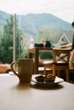 A warm café setting featuring coffee, muffins, and a scenic mountain view through the window.