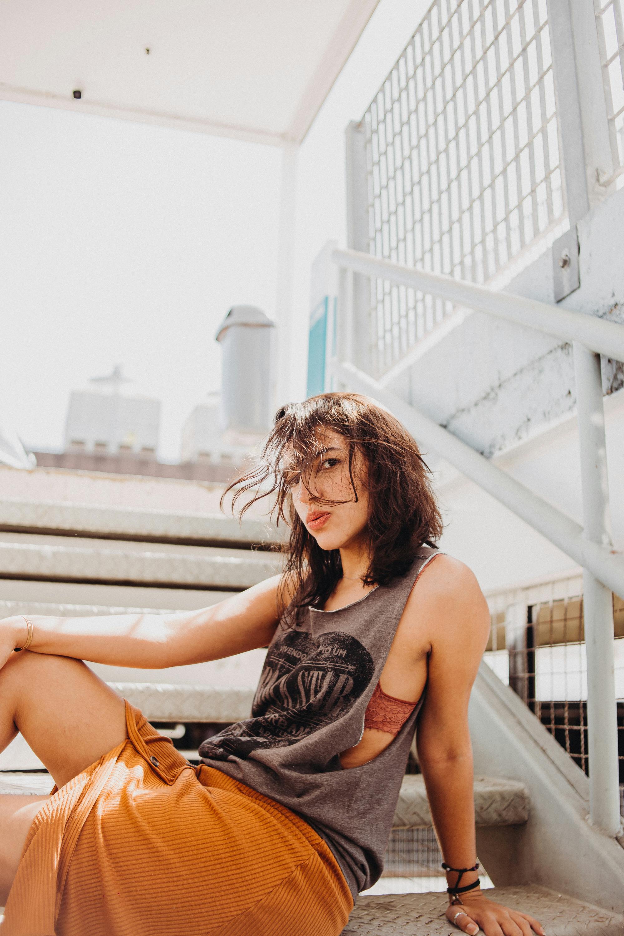 Young woman in casual clothing sitting on outdoor metal stairs, capturing an urban vibe in bright daylight.
