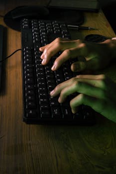 Hands typing on a computer keyboard in low lighting, capturing a focused working atmosphere.