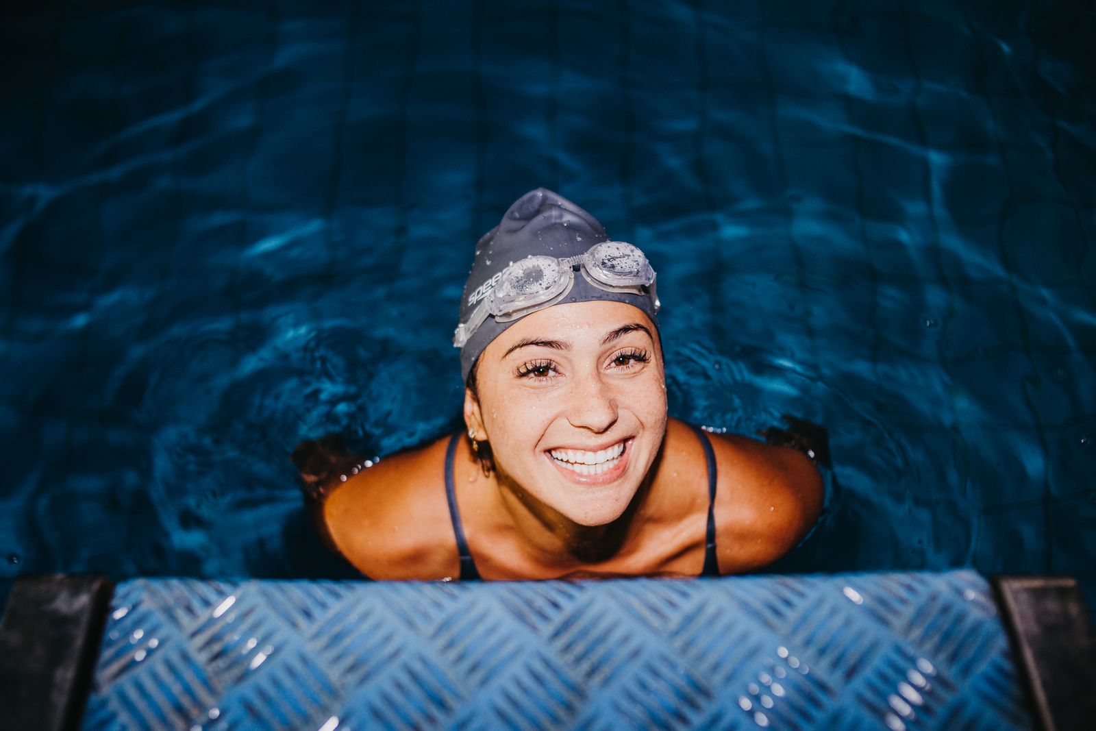 A smiling swimmer in a cap and goggles at the edge of the water