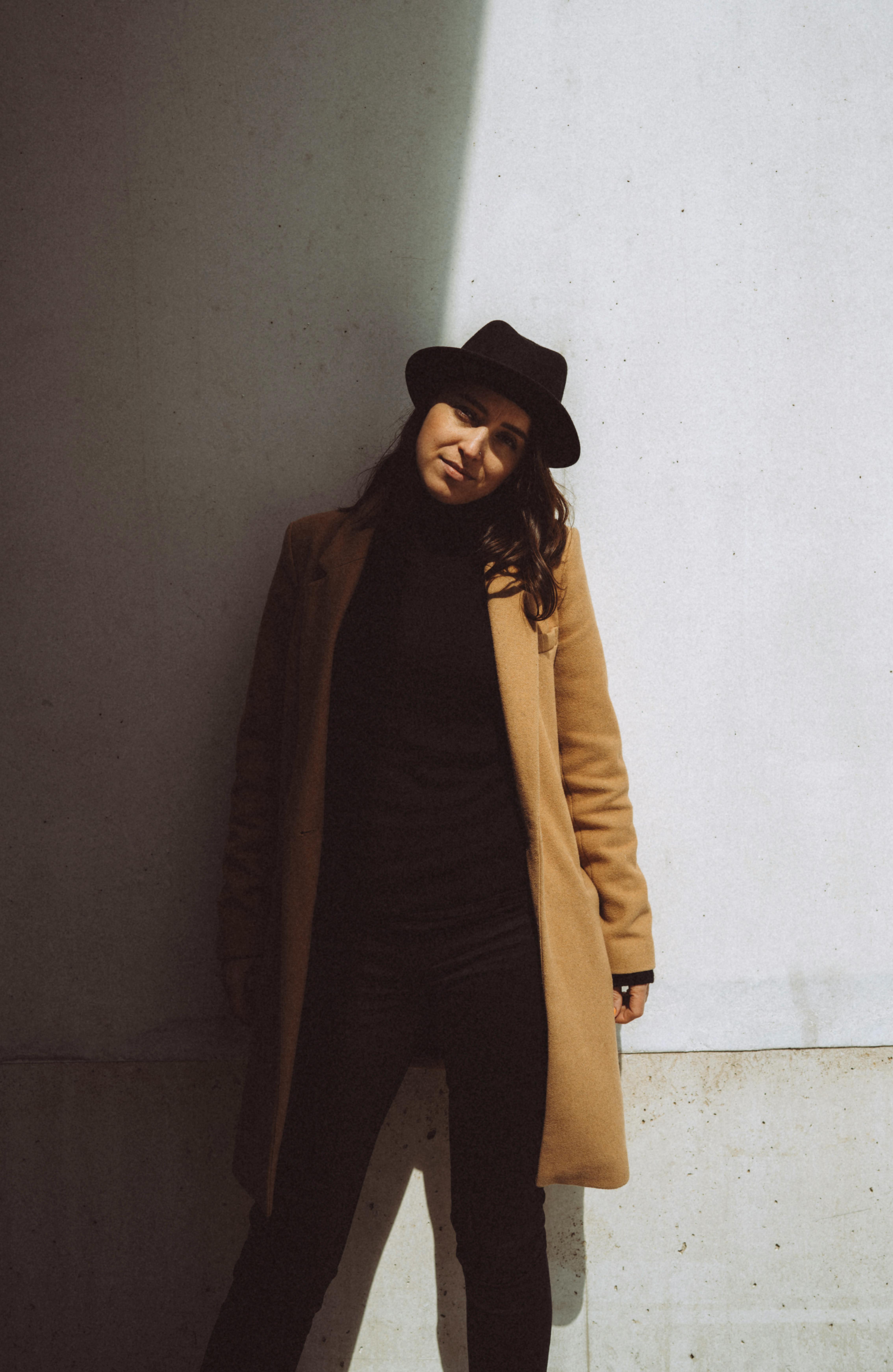 A fashionable woman stands confidently in a brown coat and black hat against a sunlit wall.