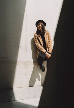 Stylish woman in a brown coat and hat posing against a sunlit wall, exuding urban chic.