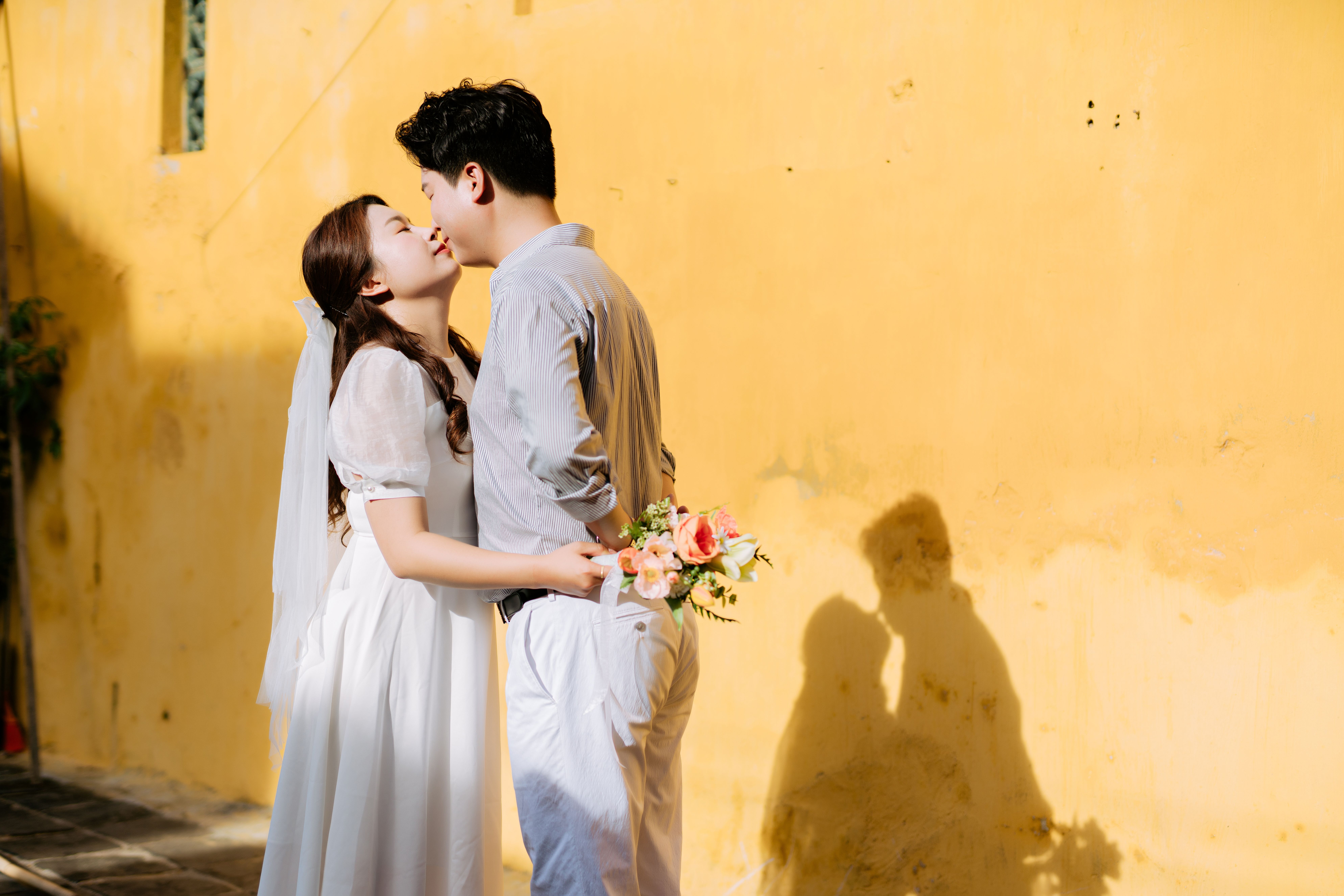 Bride and groom share a tender kiss, casting romantic shadows on a yellow wall.