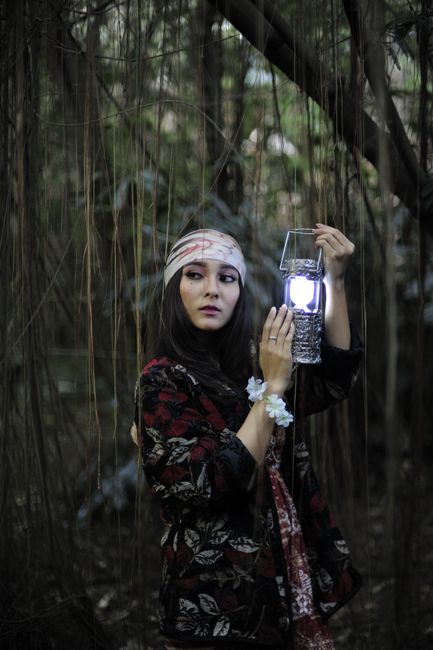 A woman in a bandana holds a lantern in a dense forest, exuding mystery.