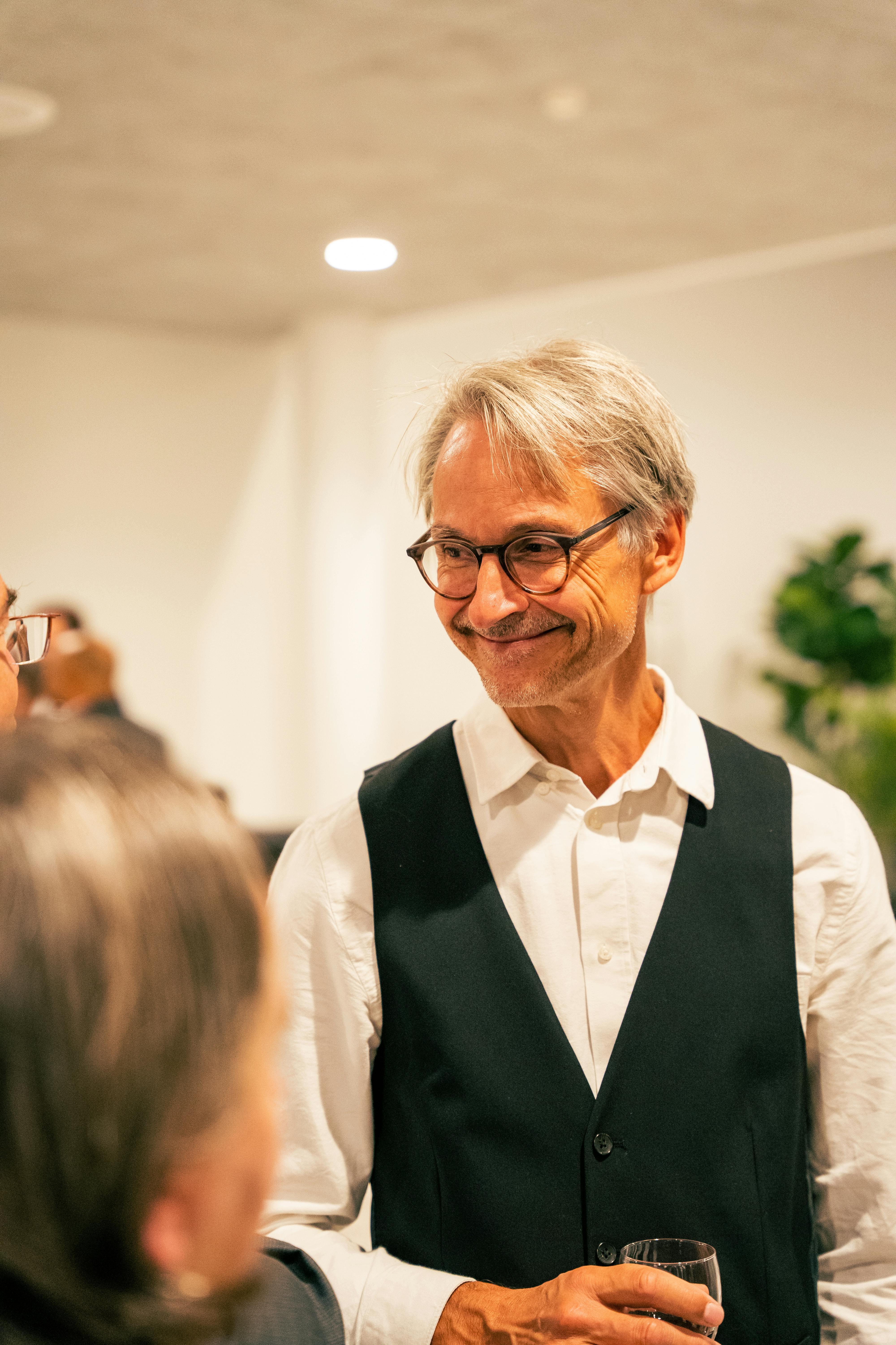 A senior man wearing eyeglasses and a vest smiles warmly at a social gathering indoors.