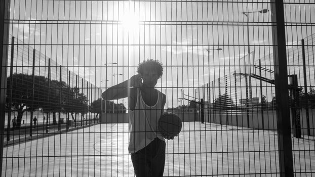 A man holding a basketball in an urban outdoor court during sunset, behind a wire fence.