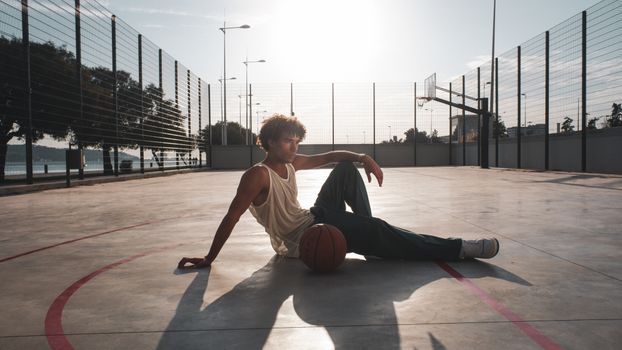 A young man sits on an urban basketball court with a ball in the warm sunlight.