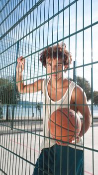 A young man holding a basketball stands by an outdoor court fence under a clear day sky.