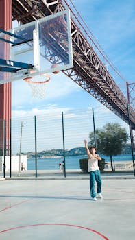 Man shoots basketball on outdoor court near iconic suspension bridge.