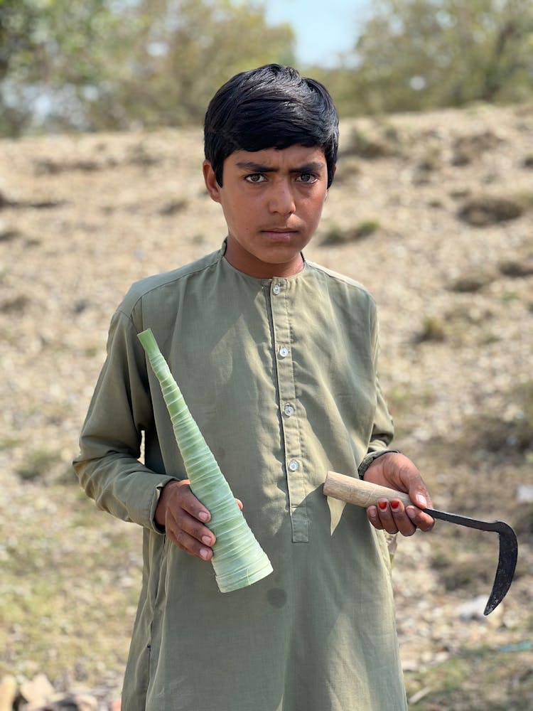 Boy In Traditional Clothes With Garden Tools In Field