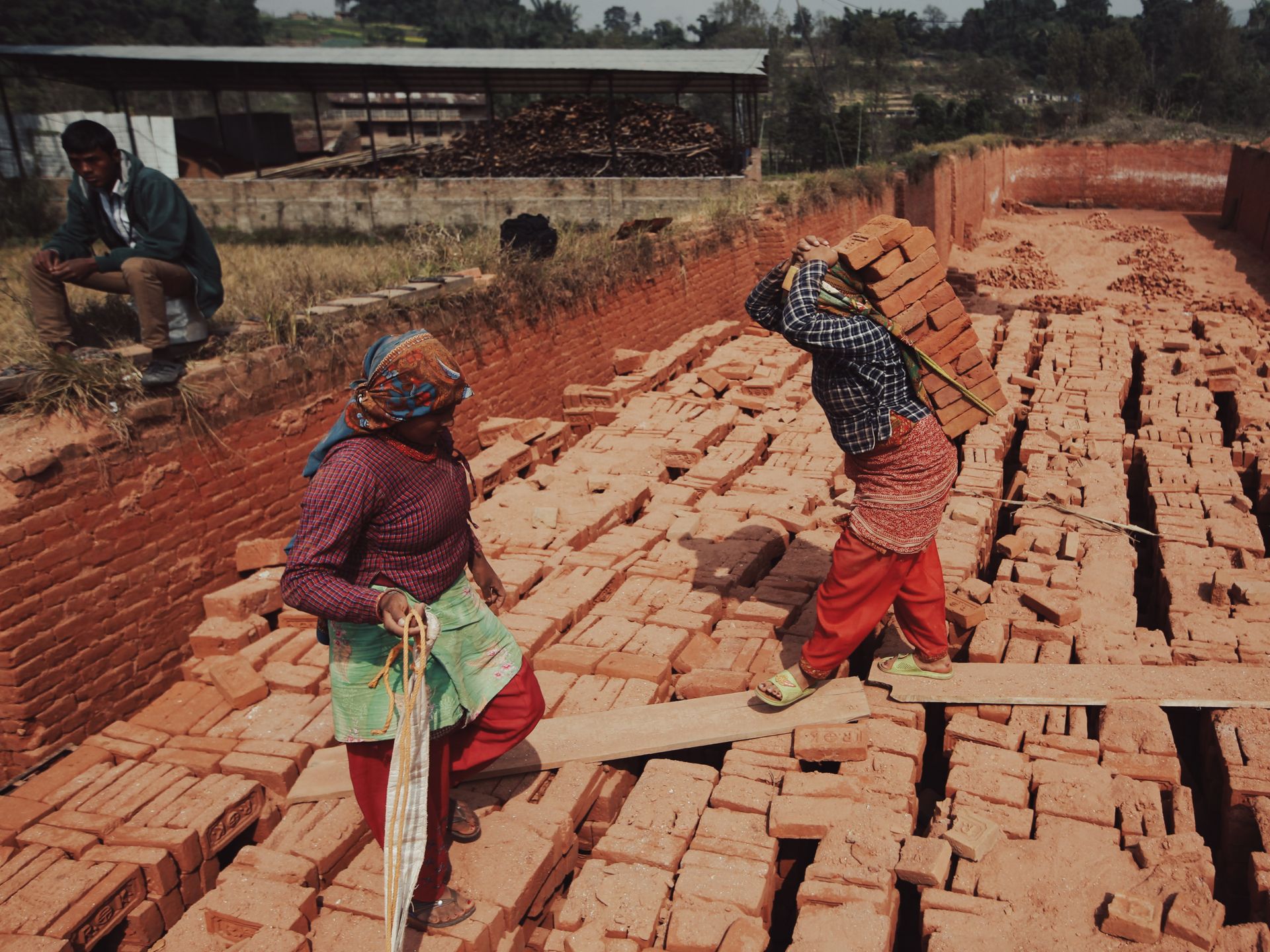 Workers engaged in manual labor carrying red bricks in an outdoor brick kiln setting during the day.