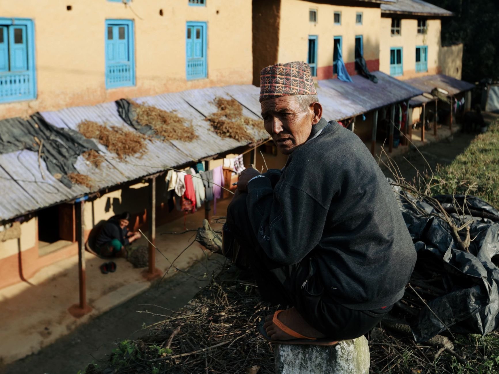 An elderly man crouches near traditional houses with blue windows in a sunlit village setting.