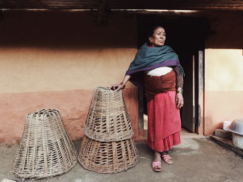 Free An elderly woman stands by woven baskets outside a rustic house, reflecting traditional lifestyle. Stock Photo