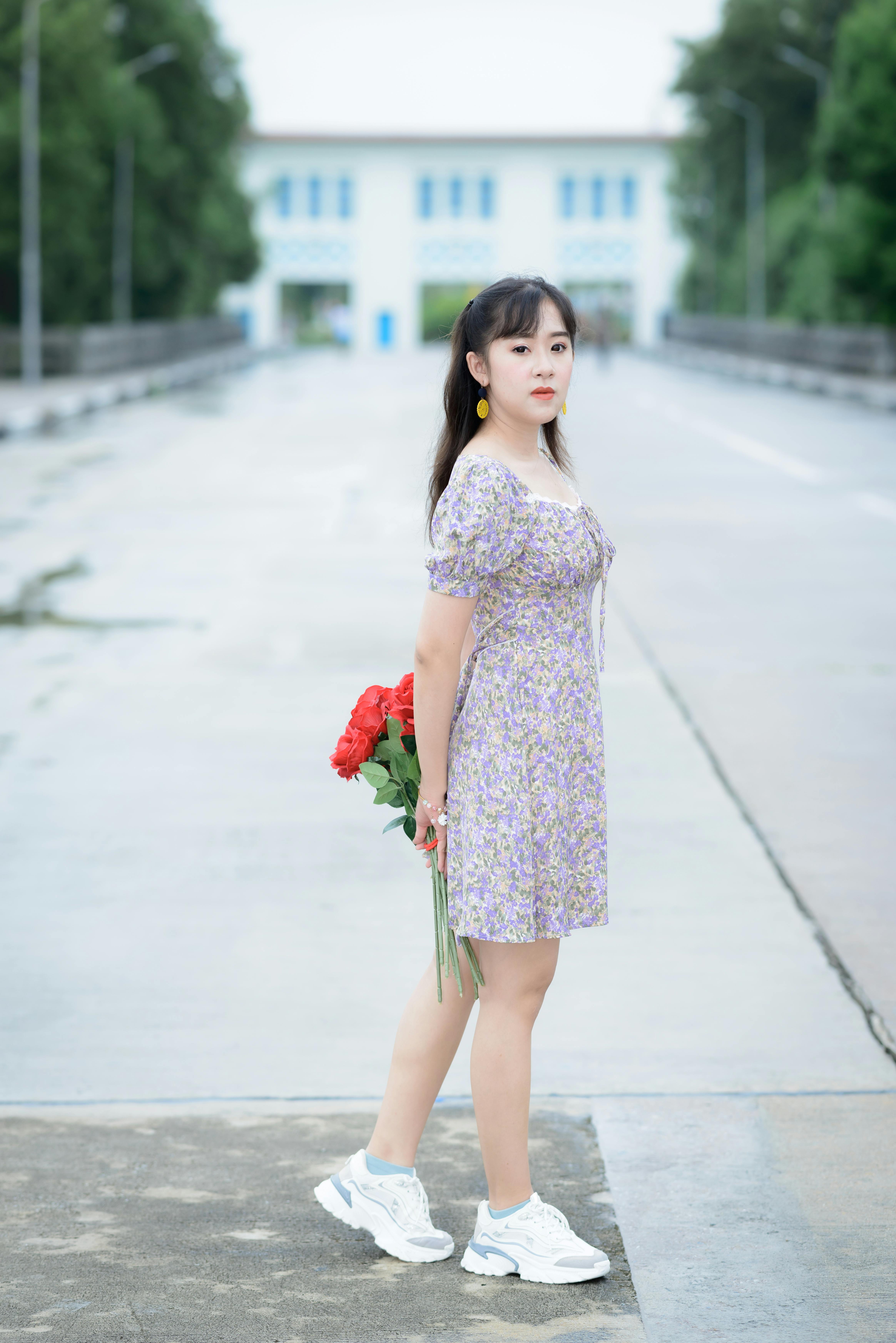 A young woman in a floral dress stands outdoors holding red roses on a clear day.