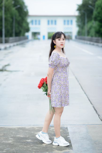 A young woman in a floral dress stands outdoors holding red roses on a clear day.