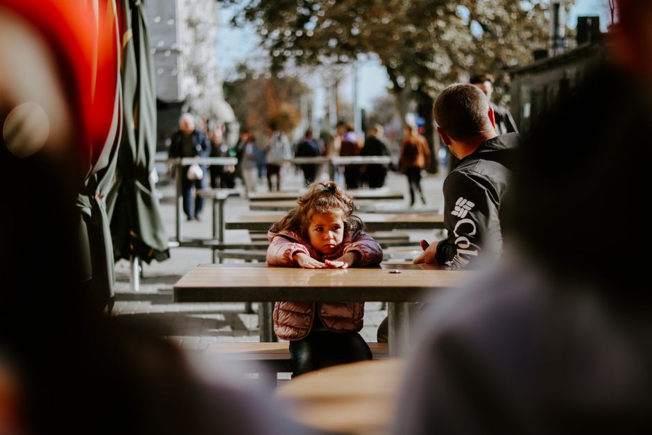 Un padre y una hija sentados en un café al aire libre mientras la gente pasa en un día soleado.
