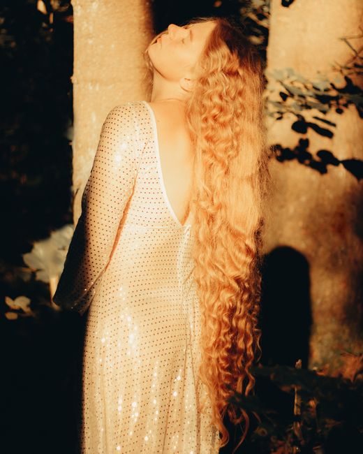 Elegant woman with long blonde hair in a glittery dress enjoying sunlight in a forest.